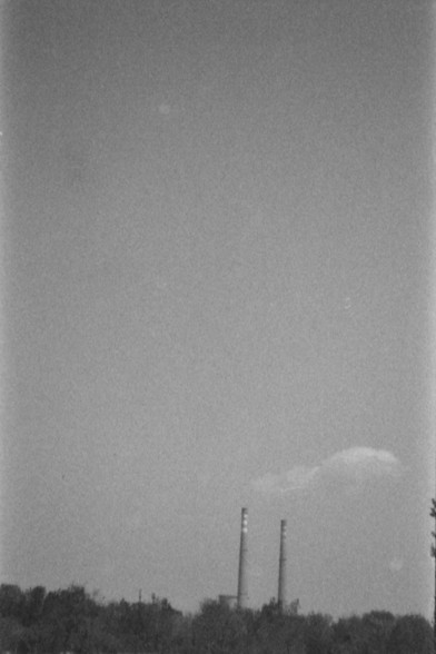 Ilford FP4 @ ISO 80

**English:**
The image is a black-and-white photograph depicting an industrial scene. In the foreground, there are trees and vegetation. Rising above the trees are two tall smokestacks, emitting plumes of smoke into the sky. The sky itself is mostly clear, with a few faint spots that could be birds or debris. The overall atmosphere suggests an industrial area, possibly a factory or power plant, set against a natural backdrop.

**Chinese:**
这张黑白照片描绘了一个工业场景。前景是树木和植被。树木上方矗立着两个…