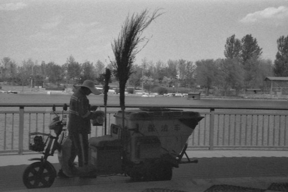 Ilford FP4 @ ISO 80

**English:**
This black-and-white photograph depicts a person standing next to a three-wheeled vehicle by a body of water. The person is wearing a hat and appears to be engaged in some activity, possibly preparing or arranging items on the vehicle. The vehicle is equipped with containers, perhaps for collecting or transporting goods. In the background, there are trees and an open area, suggesting a park or a recreational space near the water.

**Chinese:**
这张黑白照片描绘了一个人站在一辆三…