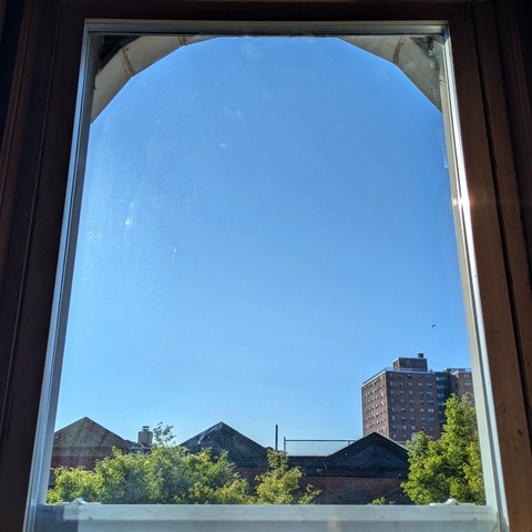 Looking through an arched window two and a half hours after sunrise the sky is bright blue and cloudless. Pointed roofs of Harlem brownstones are silhouetted across the street, and a taller apartment building can be seen in the distance. The green tops of two trees are on the bottom and right. 
