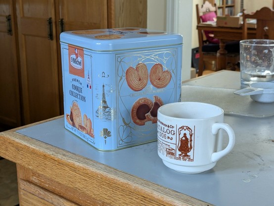 Picture of a cubic metal box of French cookies "St Michel" on the kitchen table next to a cup of coffee (not a mug) for scale.
In the background you can see a glass of water a measuring cup (1/4 of a cup) on the right. On the left there are wooden cupboard against the wall. Behind the kitchen door the wooden table of the living room.