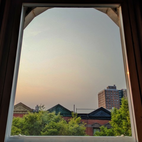 Looking through an arched window ninety minutes after sunrise the sky is a Blue of gray and peach haze. Pointed roofs of Harlem brownstones with red brickwork are across the street, and a taller apartment building can be seen in the distance. The green tops of two trees are on the bottom and right.