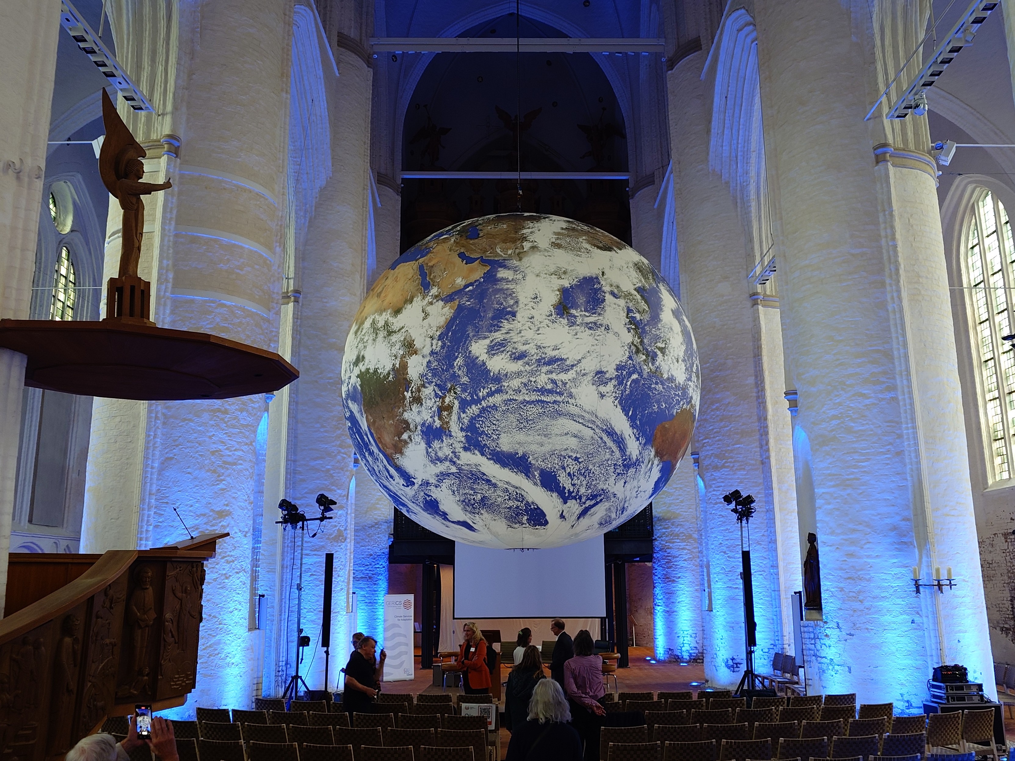 In the high vaulted main space of a large church with white columns to the sides, above the benches, hangs a huge inflated, illuminated globe of Earth with continents, oceans, clouds.