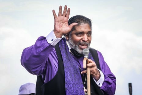 A man wearing purple liturgical garb holding a microphone and cane.