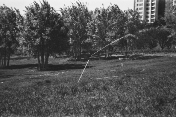Ilford FP4 @ ISO 64

**English alt text:**  
A black-and-white photograph of a large grassy field features a sprinkler system actively watering the area. The sprinkler is mounted on a tripod and sprays water in an arc, creating a fine mist that contrasts against the darker ground. The field appears well-maintained, with evenly cut grass extending across the foreground. In the background, several trees stand tall, their silhouettes adding depth to the scene. Behind the trees, a partially visible…