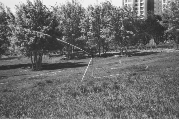 Ilford FP4 @ ISO 64

**English alt text:**  
A black-and-white photograph of a large grassy field features a sprinkler system actively watering the area. The sprinkler is mounted on a tripod and sprays water in an arc, creating a fine mist that contrasts against the darker ground. The field appears well-maintained, with evenly cut grass extending across the foreground. In the background, several trees stand tall, their silhouettes adding depth to the scene. Behind the trees, a partially visible…