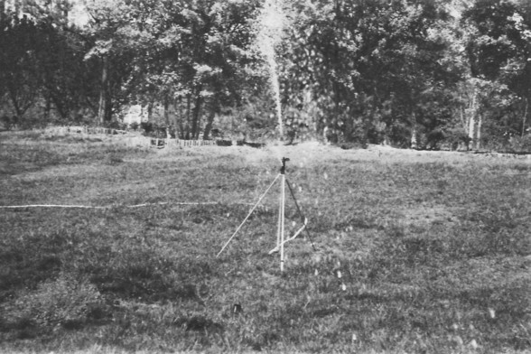 Ilford FP4 @ ISO 64

**English alt text:**  
A black-and-white photograph of a large grassy field features a sprinkler system actively watering the area. The sprinkler is mounted on a tripod and sprays water in an arc, creating a fine mist that contrasts against the darker ground. The field appears well-maintained, with evenly cut grass extending across the foreground. In the background, several trees stand tall, their silhouettes adding depth to the scene. Behind the trees, a partially visible…