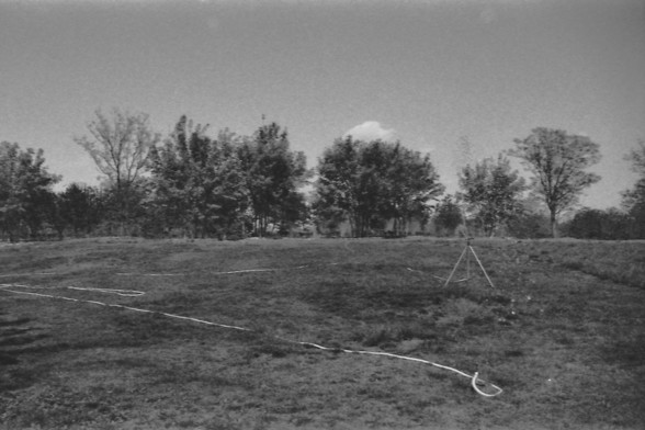 Ilford FP4 @ ISO 64

**English alt text:**  
A black-and-white photograph of a large grassy field features a sprinkler system actively watering the area. The sprinkler is mounted on a tripod and sprays water in an arc, creating a fine mist that contrasts against the darker ground. The field appears well-maintained, with evenly cut grass extending across the foreground. In the background, several trees stand tall, their silhouettes adding depth to the scene. Behind the trees, a partially visible…