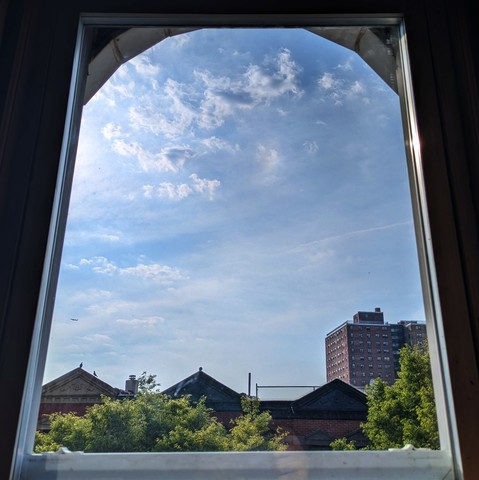 Looking through an arched window two and a half hours after sunrise the bright blue sky is covered with windswept puffy white clouds. Pointed roofs of Harlem brownstones are silhouetted across the street, and a taller apartment building can be seen in the distance. The green tops of two trees are on the bottom and right. 