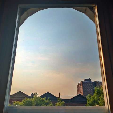 Looking through an arched window two hours after sunrise the hazy sky fades diagonally from light peach at the horizon to baby blue up above. Pointed roofs of Harlem brownstones are silhouetted across the street, and a taller apartment building can be seen in the distance. The green tops of two trees are on the bottom and right.  
