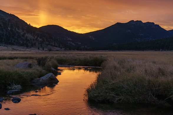 Landscape photo. Moments before sunrise in Moraine Park, in Rocky Mountain National Park. A creek flows from the bottom of the image, with low banks on either side. The creek makes a sharp left turn in the middle of the frame. Silhouetted mountains stretch across the background. The top of the frame is a deep orange with subtle cloud shapes and highlights, reflected in the creek at the bottom. October 2015.
