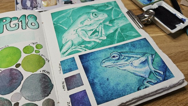 Open sketchbook spread with some swatching showing on the side, a monochromatic green frog painting, that seems to amost glow and another green and blue frog painting underneath.