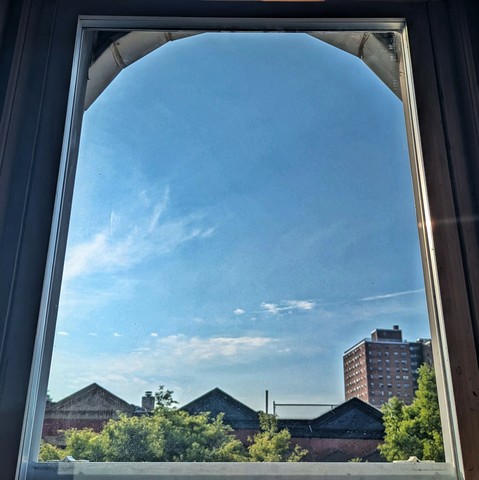 Looking through an arched window two hours after sunrise the sky is bright blue with a few scattered white clouds. Pointed roofs of Harlem brownstones are silhouetted across the street, and a taller apartment building can be seen in the distance. The green tops of two trees are on the bottom and right. 