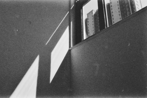 Ilford FP4 @ ISO 64

**Alt text (English):** A black and white photograph showing the interior of a room with sunlight streaming through a window, casting geometric shadows on the wall. Outside the window, tall buildings are visible.

**替代文字（中文）：** 一张黑白照片，展示了一个房间的内部，阳光透过窗户照射进来，在墙上投射出几何形状的阴影。窗外可以看到高楼大厦。
