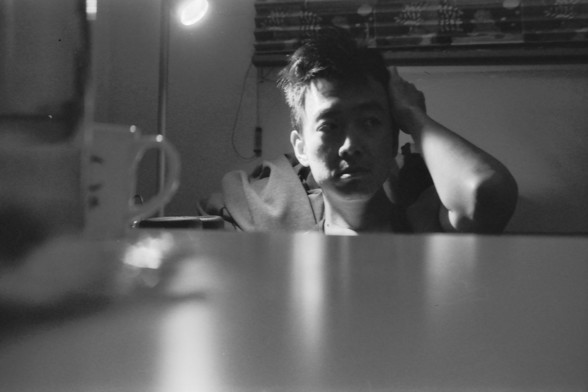 Ilford FP4 E.I. 64

**English Alt Text:**
- Description: Another black-and-white photograph of the same person, taken from a slightly different angle. The person is still seated at the table, with their head resting on their hand. The cup is visible again, and the lighting remains soft, highlighting the contours of the person's face and the texture of the table surface. The background is similarly blurred, maintaining focus on the individual.

**Chinese Alt Text:**
- 描述：另一张黑白照片，拍摄角度略有不同，但人物相同。这…