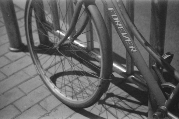 Ilford FP4 E.I. 64

**English Alt Text:**
"A black and white photograph of a bicycle parked and secured to a metal bike rack. The bike is positioned at an angle, with the front wheel prominently displayed in the foreground. The word 'FOREVER' is visible on the frame of the bicycle. The background consists of a paved sidewalk and additional bike racks."

**Chinese Alt Text:**
"一张黑白照片，展示了一辆自行车停放并固定在金属自行车架上。自行车以一定角度摆放，前轮清晰地显示在前景中。自行车车架上可以看到“FOREVER”字样。背景是铺砌的人行道和更多的自行车架。"