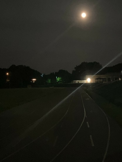 A nighttime scene featuring a running track illuminated by moonlight. In the background, there are trees and a lit building, adding a serene atmosphere. The track is clear and winding, with light reflections enhancing the nighttime view.