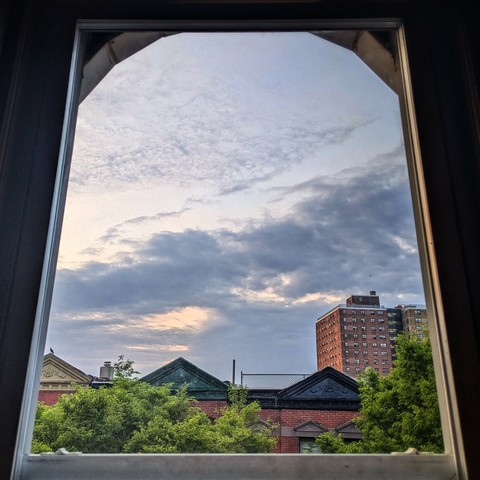 Looking through an arched window fifteen minutes after sunrise the light blue sky is crossed with thick gray-blue clouds. Pointed roofs of Harlem brownstones with red brickwork are across the street, and a taller apartment building can be seen in the distance. The green tops of two trees are on the bottom and right. 