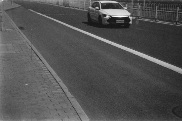 Ilford FP4 E.I. 64

English alt text: A grayscale image of a road with a white car driving toward the camera. The road is marked with a white lane line and has a brick-paved sidewalk on the left side. A fence runs parallel to the road in the background.

Chinese alt text: 一张灰度图像，展示了一条道路，一辆白色汽车正朝着镜头驶来。道路上有白色车道线，左侧是铺有砖块的人行道。背景中有一条与道路平行的围栏。