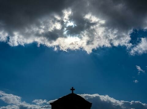 A blue sky with the sun behind a cloud. A silhouette of a cross and building visible at the bottom.
