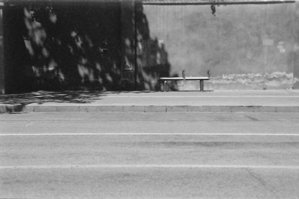 Ilford FP4 E.I. 64

**English Alt Text:**
This black-and-white photograph captures a quiet urban scene. In the foreground, there is a sidewalk and a street with visible lane markings. A bench is situated on the sidewalk, positioned against a large wall that appears to be part of a building. The wall has a textured surface and shows signs of wear and tear. The shadows of trees are cast onto the wall, adding a dynamic element to the otherwise static scene. The overall atmosphere is calm and somew…