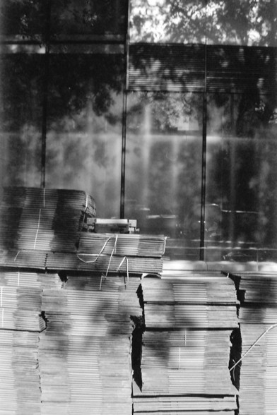 Ilford FP4 E.I. 64

**English Alt Text:**
This black-and-white photograph depicts a stack of construction materials placed in front of a modern building. The materials, which appear to be metal sheets or panels, are neatly bundled and secured with straps. The building behind the materials has a glass facade, reflecting the surrounding environment, including trees and the sky. The image captures a moment of urban development or renovation, with the contrast between the raw construction materials…