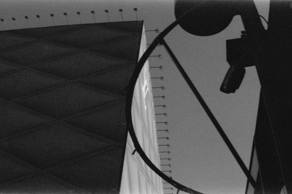 Ilford FP4 E.I. 64

**English Alt Text:**
This black-and-white image features a close-up view of a modern architectural structure. The photograph is taken from a low angle, capturing the sleek lines and geometric shapes of the building. A curved element, possibly part of a railing or decorative feature, frames the image on the right side. The building's facade is smooth and reflective, with a series of evenly spaced elements that add to its contemporary aesthetic. The contrast between light and…