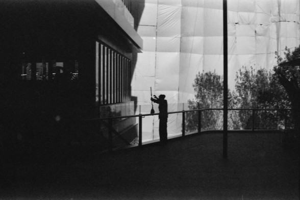 Ilford FP4 E.I. 64

**English Alt Text:**
This black-and-white image captures a person engaged in cleaning or maintenance work on a balcony of a modern building. The individual is silhouetted against the bright background, creating a stark contrast that highlights their activity. The building has a glass facade, and part of it is covered with a protective sheet, possibly for renovation or cleaning purposes. The scene is framed by the railing of the balcony, and the surrounding environment, incl…