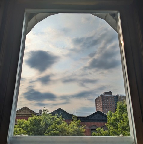 Looking through an arched window two and a half hours after sunrise the sky is full of windswept white and grey-blue clouds. Pointed roofs of Harlem brownstones are silhouetted across the street, and a taller apartment building can be seen in the distance. The green tops of two trees are on the bottom and right. 