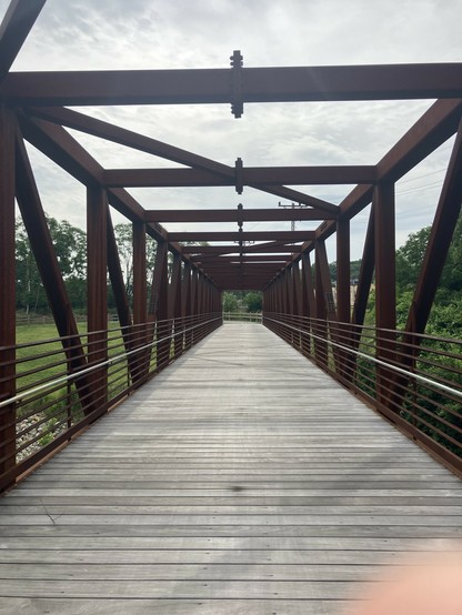 A wooden pedestrian bridge with a metal frame, surrounded by greenery and trees. The sky is partly cloudy, and the scene conveys a sense of perspective leading down the path of the bridge.