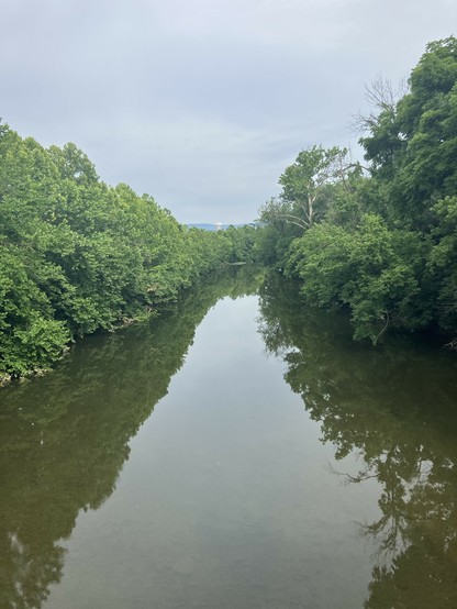A serene river reflects overhanging greenery on both banks, with a cloudy sky above and a water tower visible in the distance.