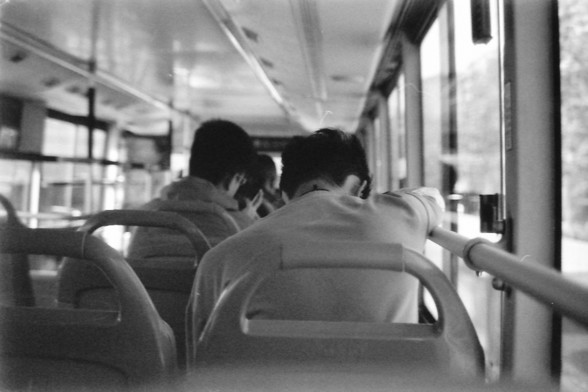Ilford FP4 E.I. 64

**English:**
This black-and-white image shows the interior of a bus. The focus is on the backs of two passengers seated near the front. The bus is in motion, as suggested by the blurred scenery visible through the windows. The interior lighting is bright, illuminating the passengers and the details of the bus seats and handrails. The image conveys a sense of movement and daily commuting.

**Chinese:**
这张黑白图片展示了公交车内部的场景。焦点集中在靠近前部的两名乘客的背影上。公交车正在行驶中，从窗外模糊的景色可以看出这一点。车内的灯光明亮，照亮了乘…