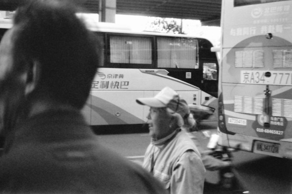 Ilford FP4 E.I. 64

**English:**
This black-and-white image captures a moment at a bus station. In the foreground, a woman wearing a cap is seen from the side, possibly talking or looking at something off-camera. Behind him, a large bus is parked, with visible text on its side. The bus station appears busy, with other people and vehicles in the background. The scene suggests a bustling transportation hub with people in transit.

**Chinese:**
这张黑白图片捕捉了公交车站的一个瞬间。前景中，一个戴帽子的女人从侧面可见，可能正在说话或看着镜头外的某物。…