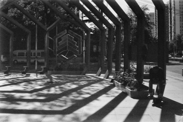 Ilford FP4 E.I. 64

English alt text: A black and white photograph of an outdoor structure with large, angled beams creating a geometric pattern of shadows on the ground. A person is sitting on a bench to the right, partially obscured by shadows. In the background, there is a bus and some buildings.

Chinese alt text: 一张黑白照片，展示了一个户外结构，带有大型倾斜梁，在地面上形成几何图案的阴影。右边有一个人坐在长椅上，部分被阴影遮挡。背景中有一辆公交车和一些建筑物。