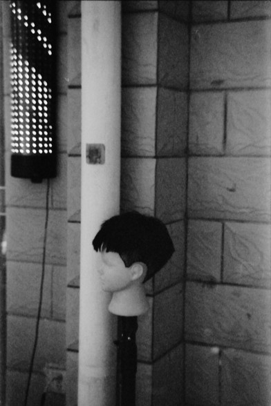 Ilford FP4 E.I. 64

English alt text: A black and white photograph featuring a mannequin head with short dark hair, placed on a stand. To its side, there is a white cylindrical object with a small rectangular sticker. Another cylindrical object with a perforated surface emits light, creating a contrast with the dark surroundings. The background consists of textured tiled walls, adding depth to the composition.

Chinese alt text: 一张黑白照片，展示了一个放在支架上的短发人头模特。旁边有一个白色圆柱形物体，上面贴着一个小矩形贴纸。另一个带有穿孔表面的圆柱体发出光…