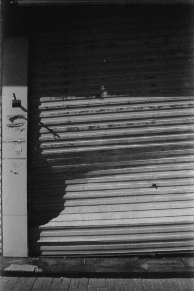 Ilford FP4 E.I. 64

English alt text: A corrugated metal shutter door with horizontal ridges, showing signs of wear and rust. A shadow is cast on the shutter from an unseen object or structure to the left. To the left of the door, a vertical column has markings or graffiti, and the ground in front is paved with tiles.

Chinese alt text: 一个带有水平脊纹的波纹金属卷帘门，表面有磨损和锈迹。左侧的未见物体或结构在门上投射出阴影。门左侧有一根立柱，上面有一些标记或涂鸦，门前的地面铺设着瓷砖。