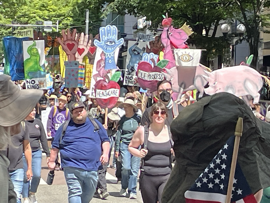 A crowd carrying colorful signs: hands in several colors, a crowned pink pig, apples with labels like “Healthcare” that the pig is eating.