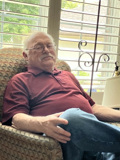 My dad, an older man with glasses and a mustache, sits comfortably in a patterned armchair, wearing a burgundy polo shirt and blue pants. Sunlight filters through window blinds behind him, highlighting the cozy indoor setting.