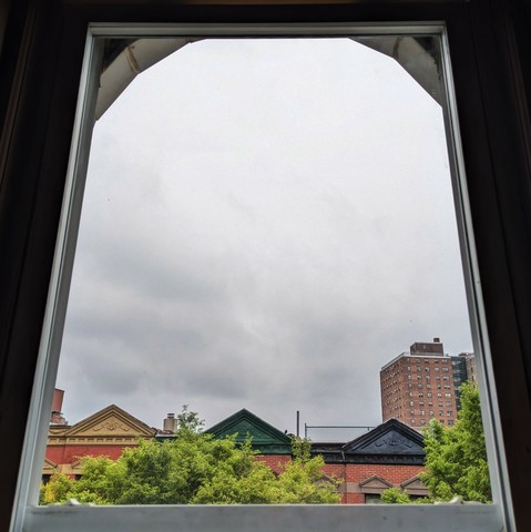Looking through an arched window two-and-a-half hours after sunrise the sky is full of grey rain clouds. Pointed roofs of Harlem brownstones with red brickwork are across the street, and a taller apartment building can be seen in the distance. The green tops of two trees are on the bottom and right.