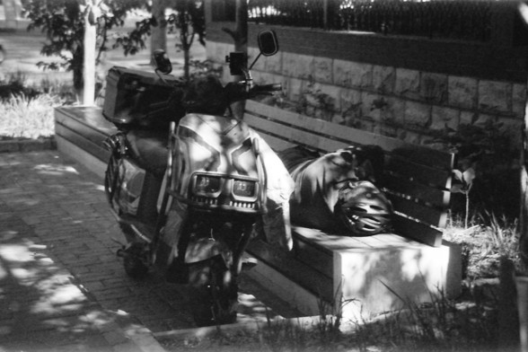 Ilford FP4 E.I. 64

English alt text: A black and white image of a parked scooter next to a bench. On the bench, there is a person lying down, wearing a helmet. The scene is shaded by trees, and there is a stone wall in the background.

Chinese alt text: 一张黑白照片，显示一辆停在长椅旁边的踏板车。长椅上躺着一个戴着头盔的人。场景被树木遮蔽，背景有一堵石墙。