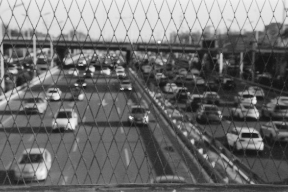 Ilford FP4 E.I. 64

English alt text: A black and white image of a busy highway seen through a diamond-patterned wire fence. The focus is on the fence, making the traffic congestion in the background appear blurred. Multiple cars occupy different lanes, emphasizing the density of urban travel.

Chinese alt text: 一张黑白照片，显示透过菱形铁丝网的繁忙高速公路。焦点集中在铁丝网上，使背景中的拥堵交通显得模糊。多辆汽车占据不同车道，突出城市交通的密集程度。