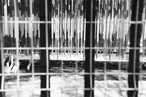 **English:**
A black and white photograph showing a close-up view of a fence made of vertical metal bars. The fence appears to be part of an industrial or construction site, with various structural elements visible in the background.

**Chinese:**
一张黑白照片，特写展示了由垂直金属条组成的栅栏。这个栅栏看起来是工业或建筑工地的一部分，背景中可以看到各种结构元素。