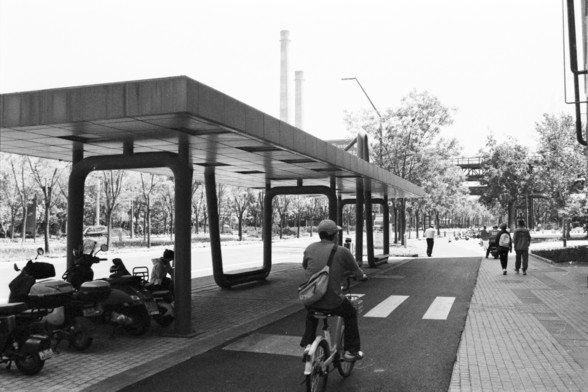 **English:**
This black-and-white photograph shows a modern urban scene with a sheltered area for scooters and bicycles. The shelter has a flat roof supported by uniquely shaped pillars. Several scooters are parked under the shelter, and a person is seen riding a bicycle on the adjacent path. The background features trees and a few pedestrians, suggesting a park or recreational area. The image captures a moment of everyday life in a city setting.

**Chinese:**
这张黑白照片展示了一个现代城市场景，其中有一个为摩托车和自行车提供的…
