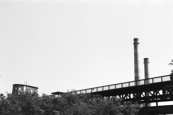 **English:**
This black-and-white photograph captures an industrial scene featuring a bridge-like structure elevated on pillars. The structure extends horizontally across the image, leading the eye towards two tall smokestacks in the background. The smokestacks are part of an industrial facility, hinting at a factory or plant. The foreground is filled with dense foliage, providing a stark contrast to the industrial elements. The sky is clear, adding to the sharpness and contrast of the image.

…