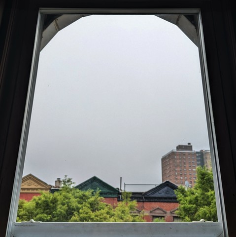 Looking through an arched window two and a half hours after sunrise the sky is full of gray clouds again. Pointed roofs of Harlem brownstones are silhouetted across the street, and a taller apartment building can be seen in the distance. The green tops of two trees are on the bottom and right. 