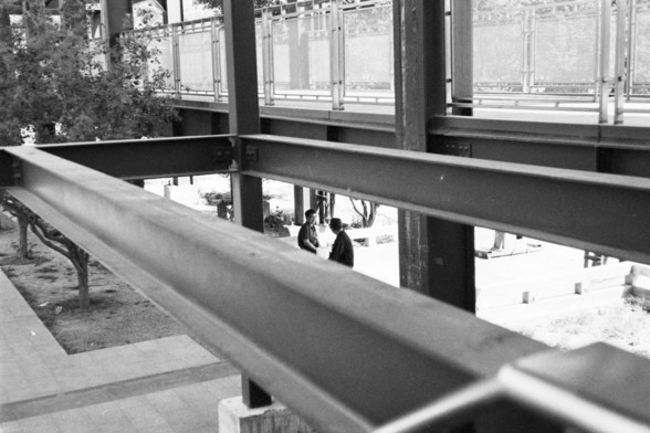 English alt text: A black and white photograph showing two people sitting and conversing under a structure with large steel beams. Above them is a walkway with railings. Trees and a paved area are visible in the background.

Chinese alt text: 一张黑白照片，显示两个人坐在一个有大钢梁的结构下交谈。他们上方是一个有栏杆的走道。背景中可以看到树木和铺砌的区域。