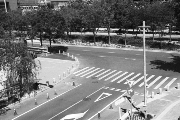 English alt text: A street intersection featuring road markings, signs, and pedestrian pathways. There is a crosswalk with white stripes, a yield sign, and a right-turn sign. Traffic cones and bollards separate pedestrian areas from the road. Trees and a building are visible in the background.

Chinese alt text: 一个街道交叉口，带有道路标记、标志和人行道。白色条纹的人行横道、让行标志和右转标志清晰可见。交通锥和路障将人行区域与道路分隔开。背景中有树木和一座建筑。