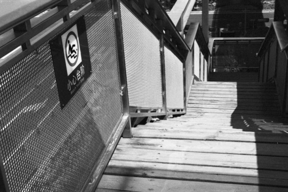 English alt text: A black and white photograph of an outdoor wooden staircase with metal railings on both sides. A warning sign attached to the left railing displays a pictogram of a person slipping, along with Chinese text “小心台阶” and English text “Watch Your Step.” Shadows partially cover the lower steps.

Chinese alt text: 一张黑白照片，显示一个户外木制楼梯，两侧有金属栏杆。左侧栏杆上附有警示牌，展示一个人滑倒的图示，并附有中文文本“小心台阶”和英文文本“Watch Your Step”。阴影部分覆盖了底部的台阶。
