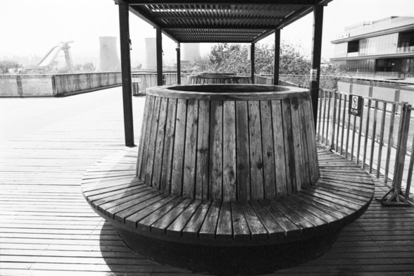 English alt text: A black and white photograph of a wooden boardwalk area featuring a circular wooden bench with a hollow center, covered by a slatted pergola-like structure. In the background, industrial buildings and cooling towers are visible, suggesting an urban or industrial setting. A sign is present on the right side, though its text is unclear.

Chinese alt text: 一张黑白照片，展示一个木制栈道区域，带有一个中空的圆形木制长椅，上方覆盖着类似凉亭的条状结构。背景中可以看到工业建筑和冷却塔，表明其处于城市或工业环境中。右侧有一个标牌，但其文字不清晰。
