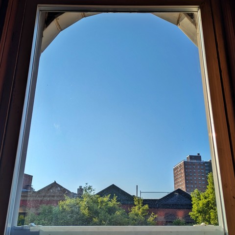 Looking through an arched window ninety minuty after sunrise the cloudless sky is bright blue. Pointed roofs of Harlem brownstones with red brickwork are across the street, and a taller apartment building can be seen in the distance. The green tops of two trees are on the bottom and right. 