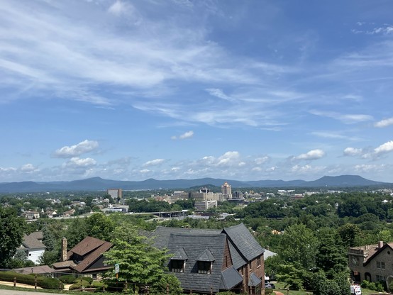 A sweeping view from partway up Mill Mountain, looking out across downtown Roanoke. Green hills comprise the foreground, covered by a blue sky with scattered clouds. Distant mountains form the northwestern edge of the valley. 